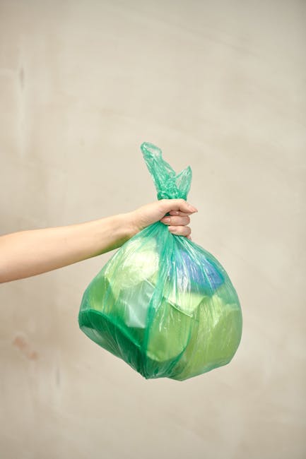 A person's arm extending into the frame from the left, holding a green and blue plastic rubbish bag tightly by the tied knot at the top. The bag appears to be filled with waste, containing a mixture of light and dark green materials, and has a slightly crumpled texture. The background is a plain, beige-colored wall with no discernible features, providing a neutral environment that emphasizes the bag. The scene is well-lit, with soft, even lighting highlighting the glossy surface of the plastic. This visual exemplifies private rubbish collection practices, possibly related to alternative waste handling or on-site clearance, as promoted by waste disposal services such as Waste Disposal Acton. The focus remains on the waste bag, illustrating an aspect of the rubbish removal process associated with household or small-scale waste management in a professional context.