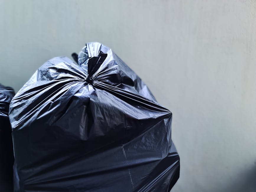 A black wheeled rubbish bin with a white label and the name 'St. John's' affixed to its side, positioned on the curbside of a quiet residential street under dim street lighting. The bin's lid is open, revealing a collection of mixed waste, including cardboard boxes and plastic bags. The street is lined with mature trees and shrubbery, casting shadows on the pavement. In the background, blurred streetlights and parked cars are visible, indicating an evening or nighttime setting. The scene suggests an alternative waste collection service, possibly provided by Waste Disposal Acton, for private rubbish disposal and on-site clearance. The pavement and street surface are well-maintained, with the bin situated just off the roadway, ready for collection or disposal of bulky waste typical of local waste management routines.