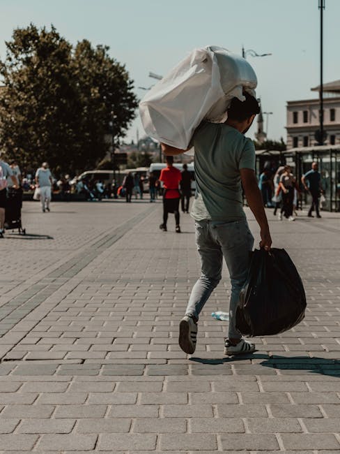 A young man walking across an open urban plaza with light grey paving stones, carrying a large white plastic bag of rubbish over his shoulder and a smaller black bag in his right hand. He wears a grey t-shirt and light grey jeans. Behind him, several pedestrians are visible, some walking and others standing, with a few engaged in conversations. In the background, there are trees, street lamps, and a mix of modern and older buildings, indicating a city environment. The scene is lit by natural daylight, casting soft shadows on the ground. The context suggests an informal scene of waste collection or disposal, possibly related to private or alternative rubbish removal services such as Waste Disposal Acton, operating within an urban setting near Acton Town station. The overall composition emphasizes the act of transporter-style rubbish handling in a public space, consistent with local waste management and rubbish removal perspectives.